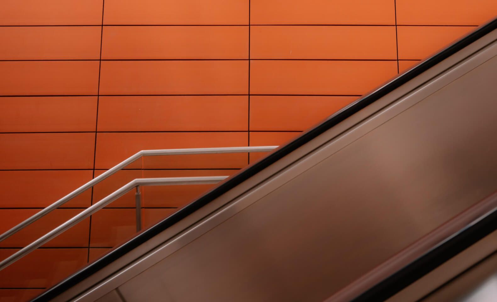 Modern escalator with sleek metal railing against an orange tiled wall – Neutrinos